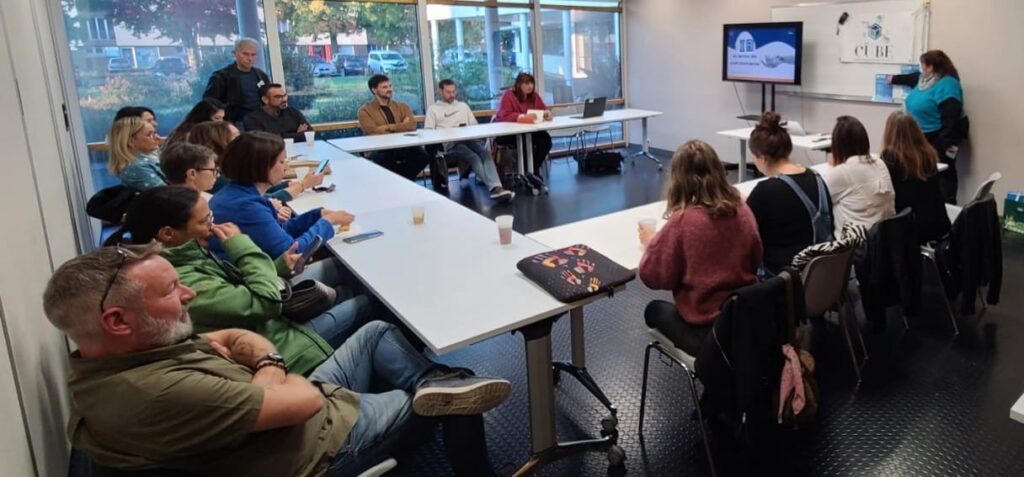 Présentation de Sandrine Crinière sur l'IA. Dans la salle en U, plusieurs entrepreneurs face à une télé qui retranscrit le powerpoint